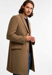 Brązowy wełniany płaszcz o dopasowanym kroju, z klapami, z zapięciem na guziki i bocznymi kieszeniami. Nosi się go na granatowym swetrze i dżinsach.