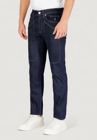 Jean en denim bleu foncé avec des coutures orange contrastantes, cinq poches et une coupe ajustée, associés à des baskets blanches.