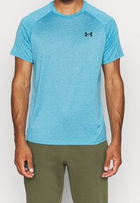 Man som bär en kortärmad blå Under Armour träningströja och olivgröna byxor, stående mot en neutral vit bakgrund.