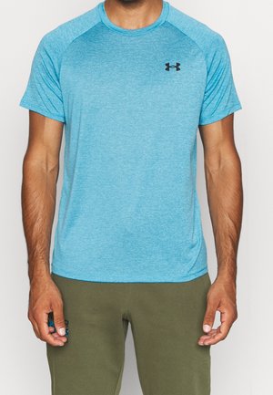 Mężczyzna w niebieskiej sportowej koszulce Under Armour z krótkim rękawem i oliwkowych spodniach, stojący na jednolitym białym tle.