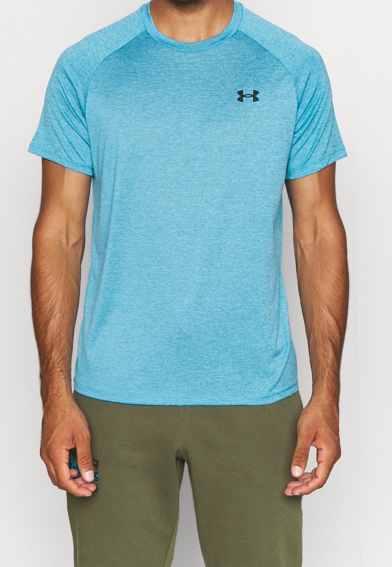 Man som bär en kortärmad blå Under Armour träningströja och olivgröna byxor, stående mot en neutral vit bakgrund.