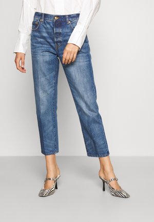 Kvinde iført blå straight-leg jeans, en hvid langærmet skjorte og zebra-mønstrede højhælede sko står på en grå gulv.
