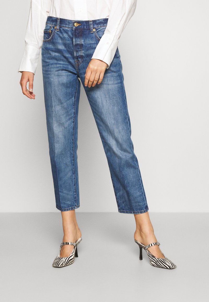 Femme portant un jean bleu coupe droite, une chemise blanche à manches longues et des chaussures à talons hauts à motif zèbre, debout sur un sol gris.