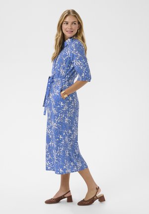 Femme debout portant une robe midi bleue à fleurs avec manches au coude, taille nouée, chaussures à talons compensés marron et bracelet en or.