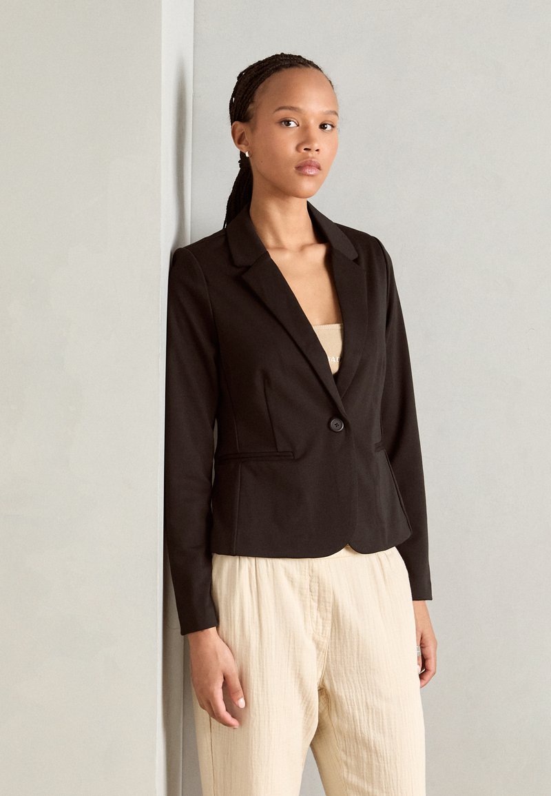Svart skräddarsydd blazer med en enkel knapp, knäppning med slag och två framfickor, bärs över en beige topp med en texturerad yta.