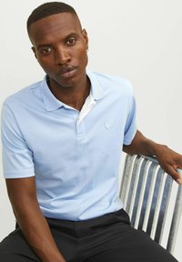 Világoskék polo felső, fehér gallérral, ami mellett egy kis logó található. Rövid ujjú, sima anyagú, sötét nadrággal párosítva.