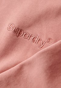 La tela de algodón rosa presenta el nombre de la marca "Superdry" bordado en una textura suave y sutil, mostrando un diseño limpio y minimalista.