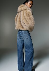 Femme portant une veste en fourrure beige et un jean bleu à jambes larges avec des poches arrière cloutées, debout devant un fond gris uni.