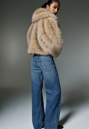Ženska, oblečena v bež krzneno jakno in široke modre kavbojke z nabranimi zadnjimi žepi, stoji na sivem ozadju.
