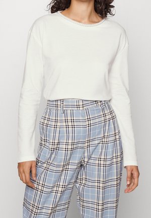 Femme portant un haut blanc à manches longues et un pantalon à carreaux bleu et beige taille haute avec des plis et des passants de ceinture.