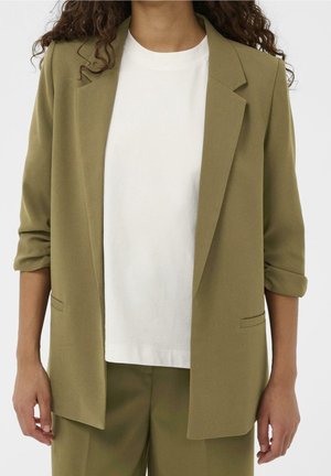 Persoon draagt een olijfgroene blazer met opgerolde mouwen over een effen wit T-shirt met ronde hals en bijpassende olijfgroene broek.