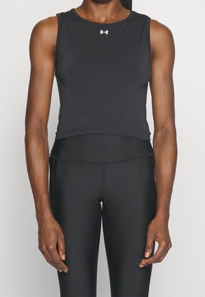 Osoba oblečená v černém sportovním tílku bez rukávů s bílým logem Under Armour a odpovídajících černých přiléhavých legínách, stojící před jednoduchým pozadím.