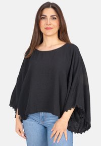 Schwarzes Poncho-Oberteil aus leichtem strukturiertem Stoff, mit weiten Ärmeln und Spitzenbesatz am Saum, kombiniert mit blauen Jeans.