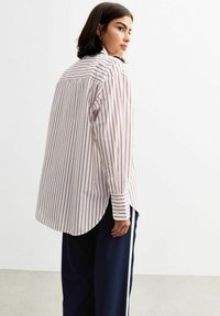 Wit langemouwen shirt met verticale rode strepen. Heeft een gebogen zoom, knoopmanchetten en een relaxte pasvorm. Draag het met donkere broeken.