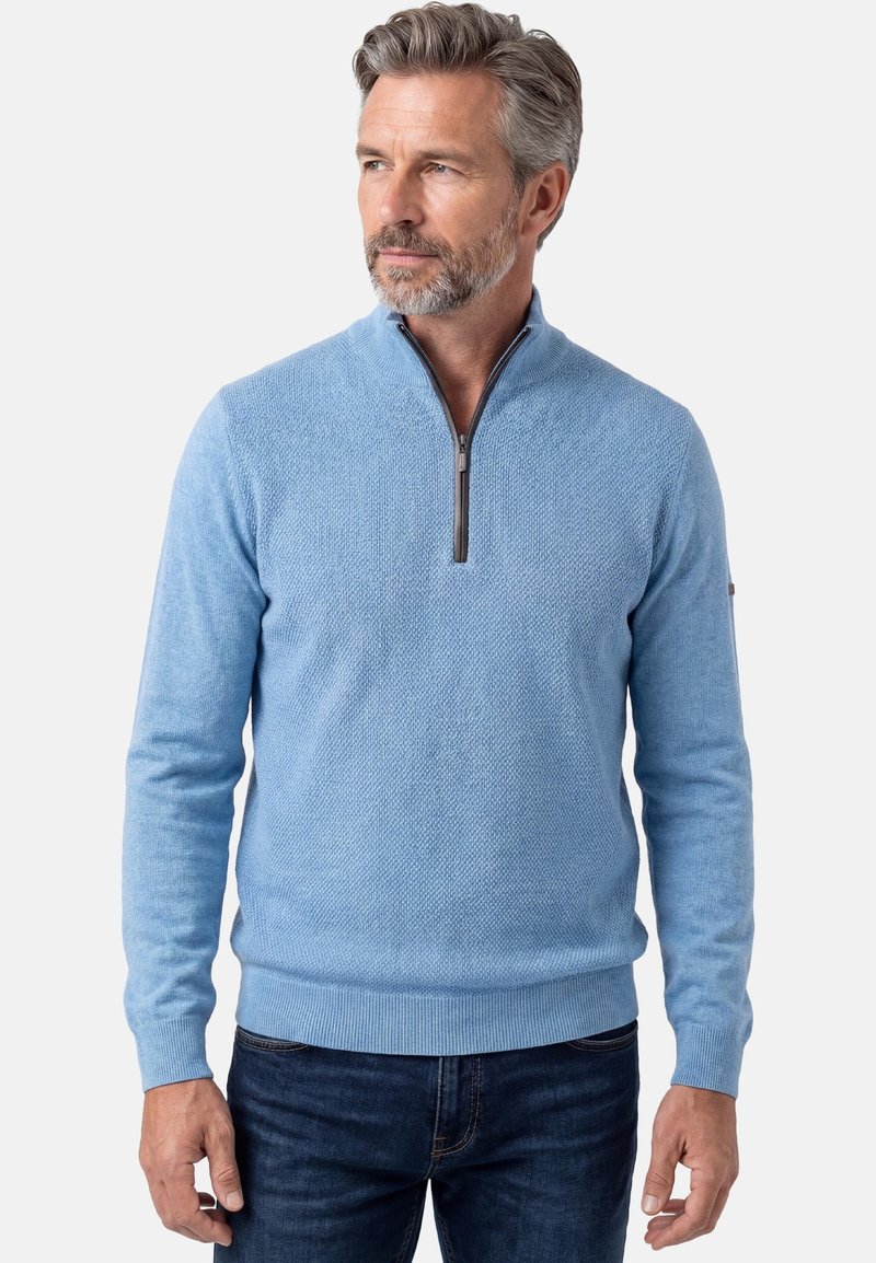 Mittelalter Mann trägt einen hellblauen, strukturierten Pullover mit Viertel-Reißverschluss und dunkle Jeans, schaut leicht nach links vor einfarbigem Hintergrund.