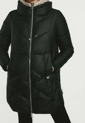 Femme portant un long manteau matelassé noir avec fermeture éclair à l'avant, capuche et mains dans les poches sur un fond clair uni.