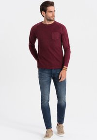 Chemise bordeaux à manches longues en maille côtelée avec une poche poitrine, associée à un jean bleu et des baskets beiges, debout devant un fond neutre.