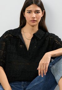 Schwarzes, häkelartiges Oberteil mit Kragen, das ein Gittermuster aus floralen Ausschnitten zeigt. Das Model trägt blaue Jeans.