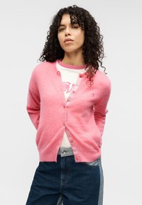 Ροζ, χνουδωτή ζακέτα με V-λαιμόκοψη, κουμπιά μπροστά και μακριά μανίκια, φορεμένη πάνω από λευκό πουκάμισο με ροζ λεπτομέρειες. Φαίνονται τζιν παντελόνια.