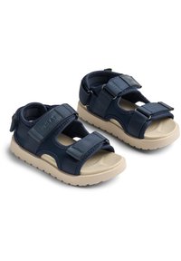 Tummansiniset sandaalit säädettävillä Velcro-hihnoilla, pehmeä kangas päällä ja vaaleanruskea kumipohja. Teksturoitu pohja lisää mukavuutta.