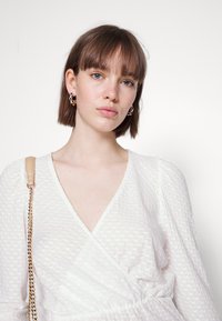 Top branco envolvente com tecido texturizado, apresentando mangas compridas e decote em V. Bolsa com alça de corrente dourada pendurada no ombro.