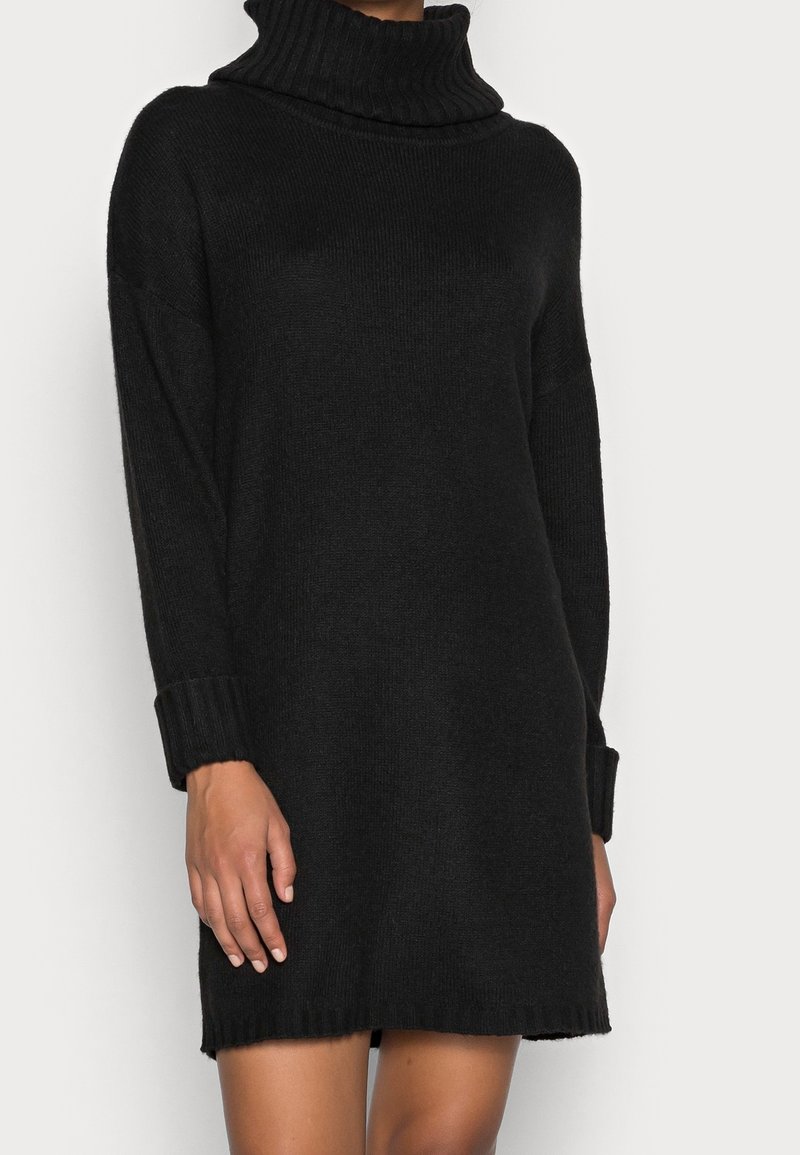 Robe-pull en maille noire à manches longues avec un col roulé haut replié, des poignets côtelés et un ourlet légèrement effiloché.