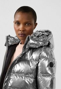 Metalen zilveren donsjack met capuchon, voorzien van een ritssluiting, elastische manchetten en een logopatch op de linkerarm.
