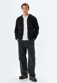 Zwarte jack met capuchon en witte stiksels, gecombineerd met losse zwarte cargobroek met zijzakken en een wit t-shirt eronder.