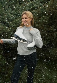 Balti slidošanas apavi ar melniem asmeņiem, ko tur rokās cilvēks, kurš valkā gaiši pelēku džemperi ar izšuvumiem un tumšas bikses, kamēr krīt sniegs.