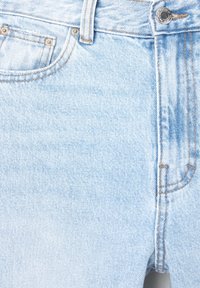 Jeans in denim azzurro, con un primo piano che mostra la tasca anteriore, il passante per cintura, i rivetti metallici e i dettagli del bottone con cuciture arancioni.
