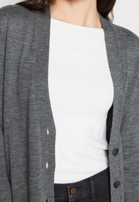 Szary kardigan o luźnym kroju, z dekoltem w kształcie litery V, z zapięciem na guziki i prążkowaną fakturą, nałożony na biały t-shirt.