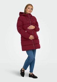 Burgundská péřová bunda s kapucí, s horizontálním prošíváním, předním zipem a bočními kapsami; v kombinaci s modrými džíny a černými botami.