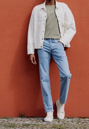 Weiße Jeansjacke, olivgrünes T-Shirt mit Logo, hellblaue Jeans und weiße Sneaker. Strukturiertes Material, lässige Passform, simples Design.