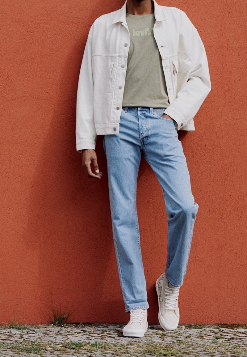 Vit denimjacka, olivgrön t-shirt med logotyp, ljusblå jeans och vita sneakers. Strukturerad tyg, avslappnad passform, enkel design.