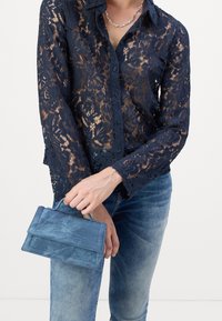 Chemise boutonnée en dentelle marine avec motif floral, manches longues, portée avec un jean en denim bleu clair et un petit sac à main bleu.