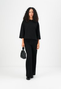 Schwarzes Cropped Top mit strukturiertem Muster, kombiniert mit weit geschnittenen schwarzen Hosen. Accessoires sind eine kleine schwarze Tasche und schwarze Schuhe.