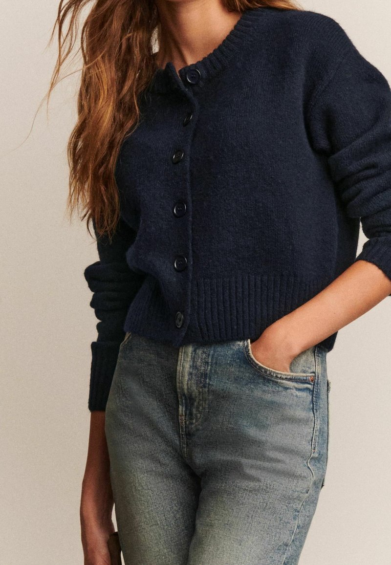 Femme portant un cardigan bleu marine foncé boutonné avec des poignets côtelés et un jean bleu clair délavé, main posée négligemment dans la poche.