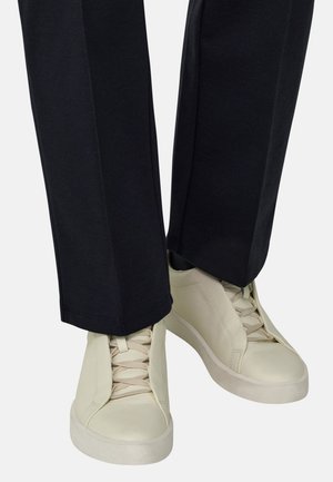 Personne portant un pantalon noir et des baskets en cuir écru avec des semelles larges et des lacets assortis, debout sur un fond clair.