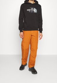 Černá mikina s kapucí s logem "The North Face", zkombinovaná s jasně oranžovými kalhotami a černými turistickými botami. Neformální venkovní oblečení.