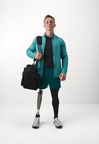 Sportiškas vyras modelis mėtų spalvos švarku ir šortais, juodomis kojinėmis, naudojantis protezine koja, laikantis juodą maišą, vilkintis pilkai sportbačiais.