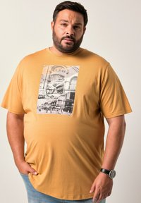 Muž s vousy, oblečený v hořčicově žlutém tričku s vintage potiskem městské krajiny, modrých džínách a černými náramkovými hodinkami, stojí na světlém pozadí.