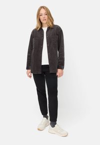 Kohlenstofffarbene Cordjacke mit zwei Vordertaschen, getragen über einem weißen Oberteil und kombiniert mit schwarzen Hosen und beigen Sneakers.