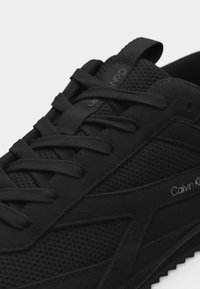 Gros plan sur une basket Calvin Klein noire avec des panneaux en mesh et en suède, des lacets plats noirs, et le logo de la marque visible sur le côté et la languette.