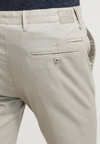 Pantaloni beige su misura con tasca a filetto sul retro con bottone e etichetta del marchio in tessuto vicino all'elastico, indossati con un top scuro e testurizzato.