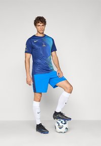 Blå Nike fodboldtrøje med grøn gradientdesign, parret med blå shorts. Sorte fodboldsko og hvide sokker. Bolden har et mønster.