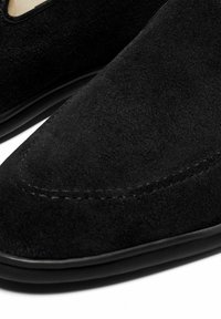 Schwarze Wildlederschuhe mit einem spitzen Zehen-Design, das gestickte Details und eine Gummisohle aufweist. Die Nahaufnahme hebt die Textur und Farbe hervor.