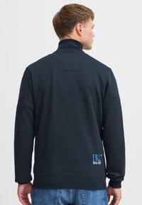 Marineblå pullover-sweatshirt med høj hals, ribbet krave og manchetter. Har et lille blåt logo på den nederste ryg. Fremstillet af bomuld, afslappet pasform.