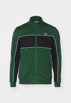 Groene ritsjack met zwarte horizontale streep, witte piping en Lacoste-logo op de linkerborst en mouw. Gemaakt van gladde stof.