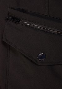 Svart tyg med en klaffficka och ett silverfärgat dragkedjestängning. Har en rund metallknappdetalj. Sl ät textur och skräddarsydd design.