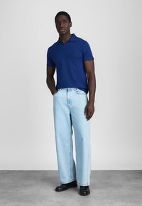 Homem de pé com as mãos nos bolsos, vestindo uma camisola polo de mangas curtas azul, jeans de perna larga azul claro e sapatos pretos, em um chão cinza.